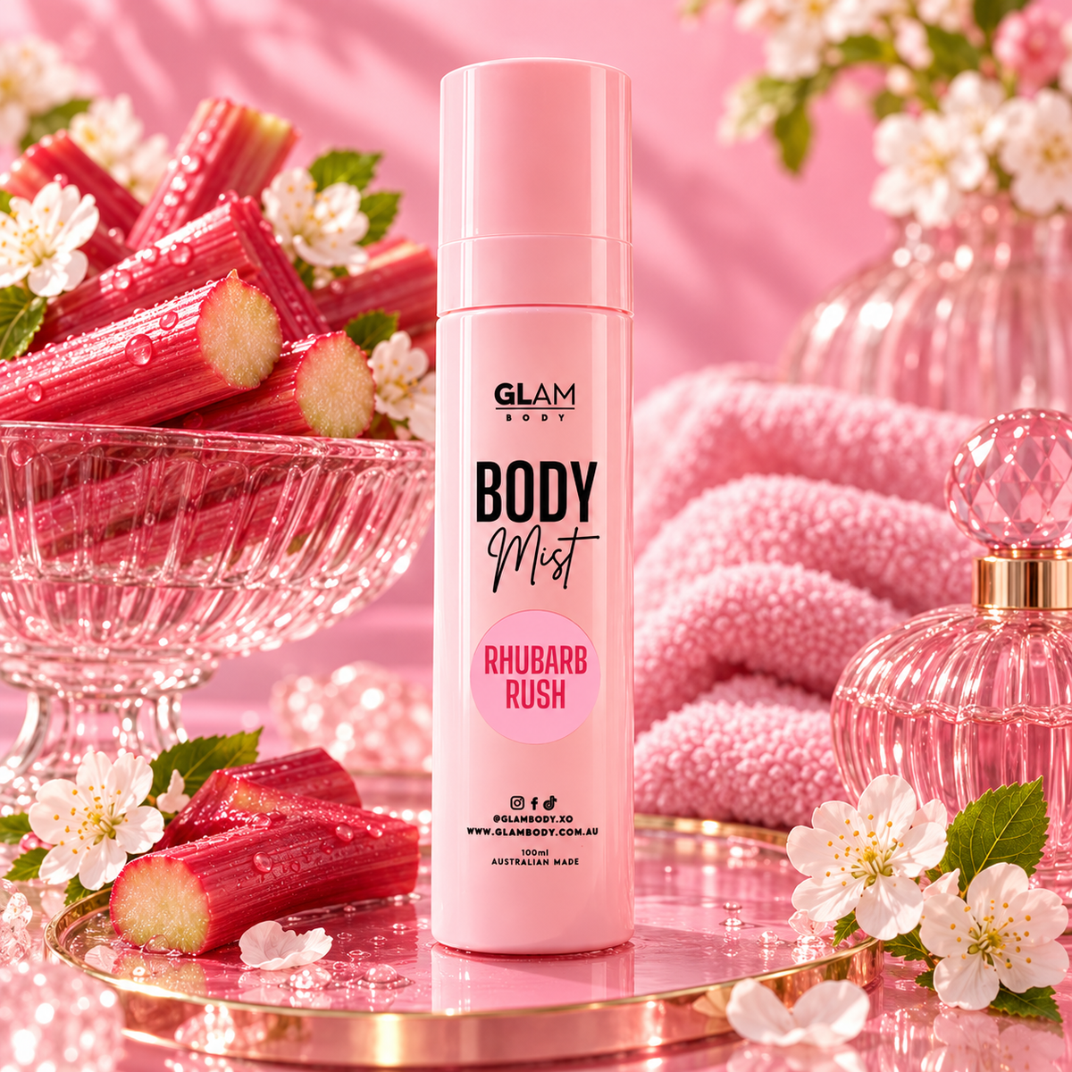 A pink cylindrical bottle of GLAM BODY Body Mist Rhubarb Rush stands on a reflective pink tray, surrounded by pink-toned rhubarb stalks, white blossoms, a pink towel, and a pink perfume bottle in a styled studio setup.