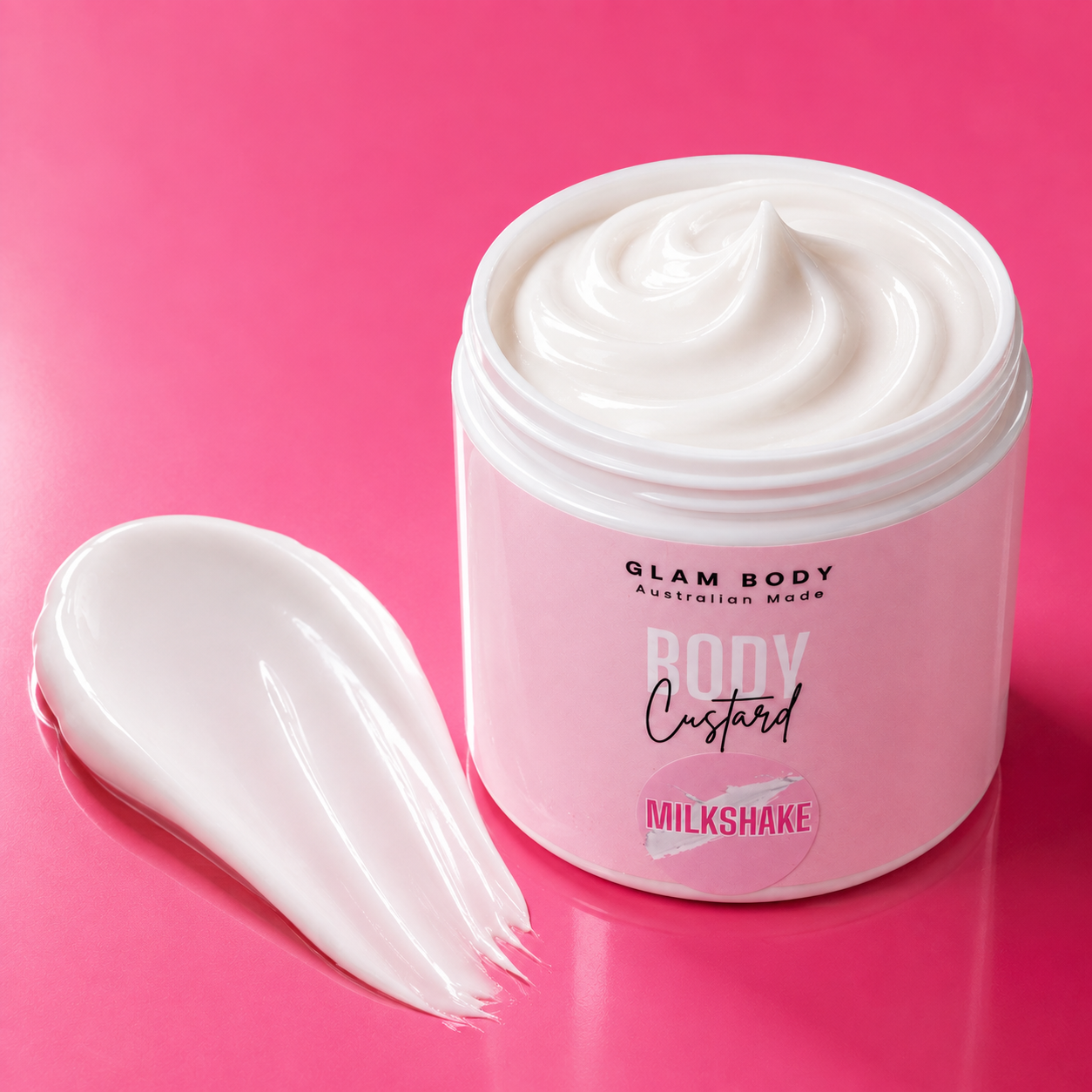 A jar of Glam Body Australian Made Body Custard in the Milkshake flavor, shown open with a swirl of thick white cream inside and a smear of the cream on a vibrant pink background.
