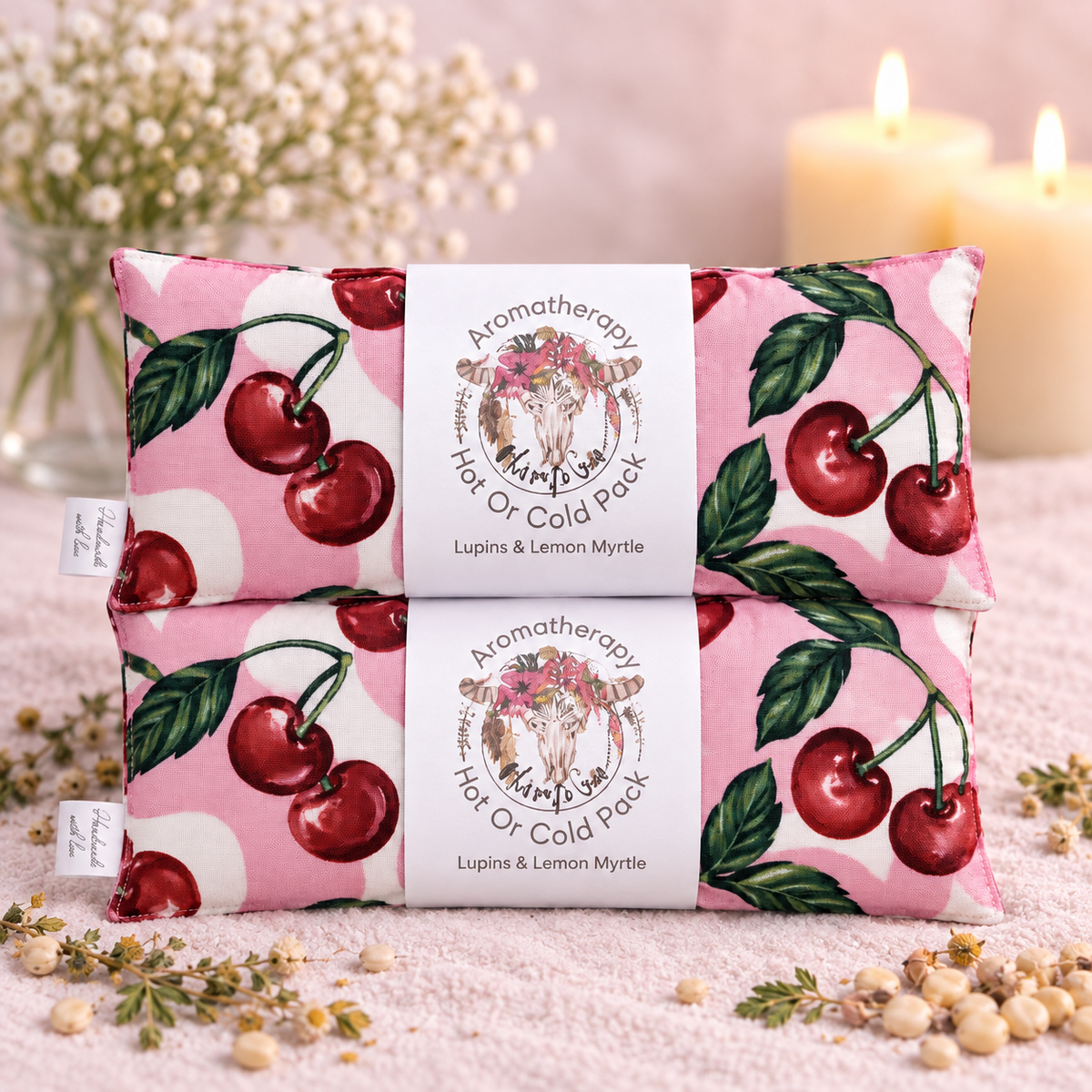 Two pink aromatherapy neck or cold packs stacked on a soft pink surface, decorated with cherry and daisy illustrations. Each pack has a white paper band label in the center reading 'Aromatherapy Happy Dog' and 'Lupins & Lemon Myrtle'.