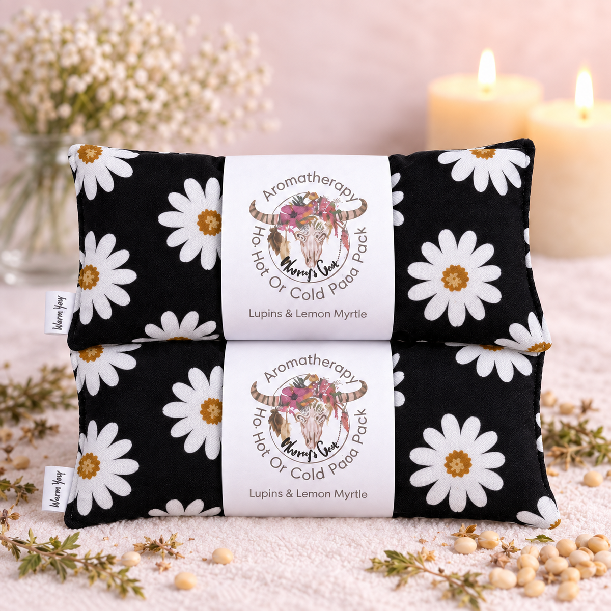 Two black fabric aromatherapy neck or cold pack pads with a white daisy print, stacked on a textured surface with dried flowers, candles, and beads. Each pad has a white paper band label in the center reading 'Aromatherapy Hair Or Cold Pack Pack' with a cow illustration and 'Lupins & Lemon Myrtle' by 'Karyn Lee'.
