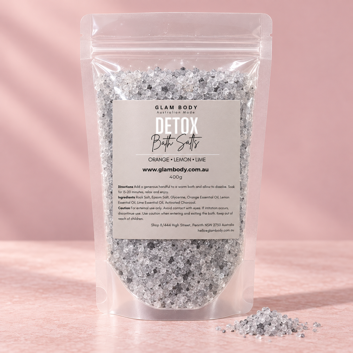 A stand-up resealable pouch of Glam Body Detox Bath Salts filled with silver-colored bath salts, set against a soft pink background with a few salts spilled in front.