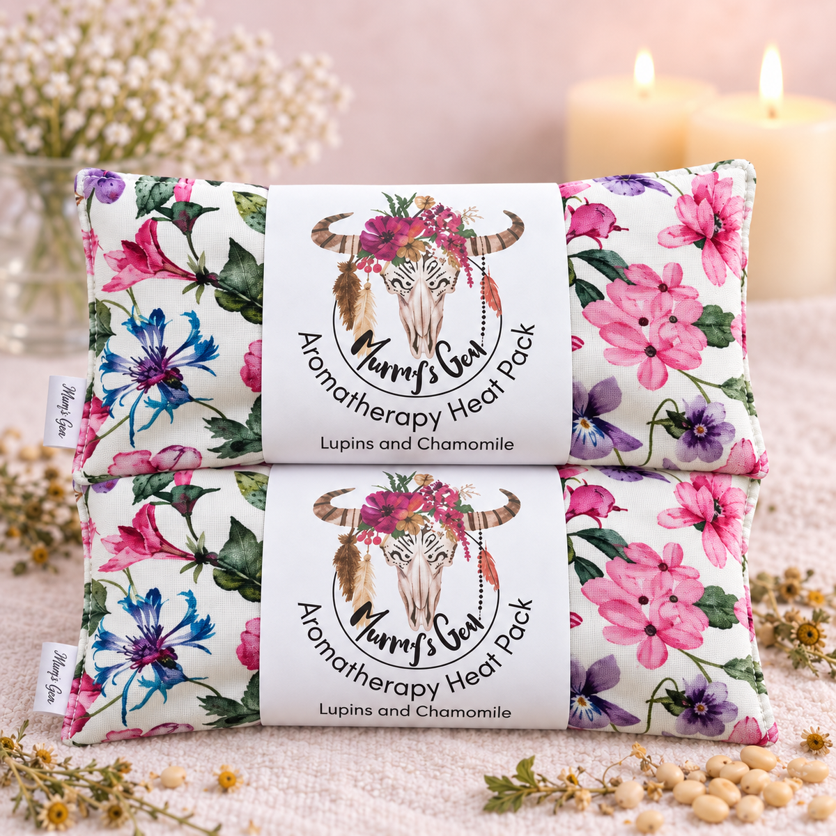 Two packaged aromatherapy heat packs stacked on a soft fabric surface, with flowers, candles, and small dried botanicals in the background. The packs have a white center label reading 'Murris Bear Aromatherapy Heat Pack' and 'Lupins and Chamomile,' with pink, purple, blue, and green floral artwork on the sides.