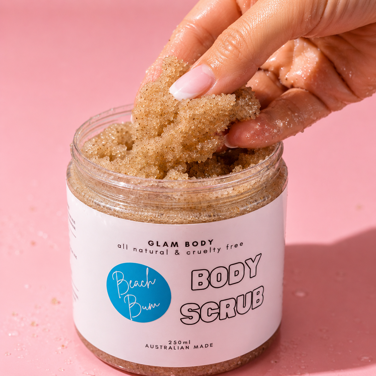 Hand scooping body scrub from a jar labeled 'Glam Body' on a pink background