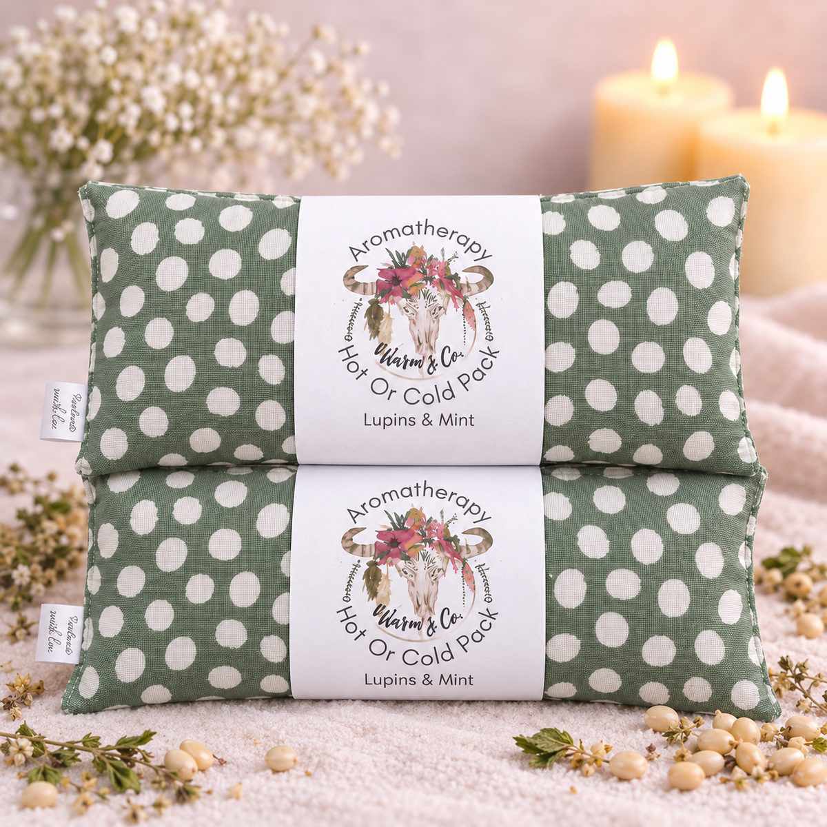 Two green and white polka dot aromatherapy hot or cold packs stacked on a soft fabric surface with dried flowers, seeds, and lit candles in the background. Each pack has a white paper band labeled 'Aromatherapy Hot or Cold Pack' with a floral bull skull illustration and the text 'Lupins & Mint'.