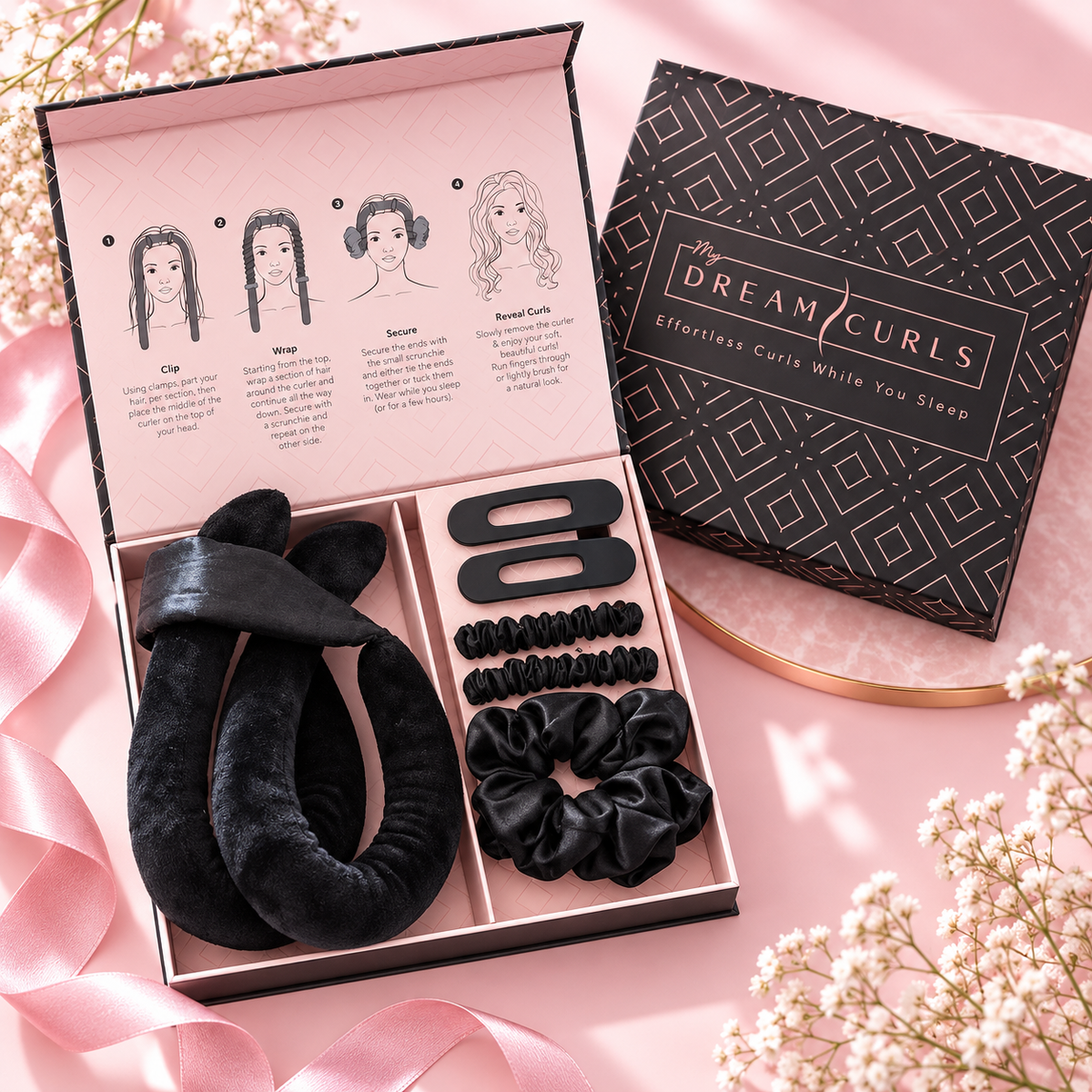 A hair styling gift set from Dream Curls, featuring black velvet scrunchies, a black hair clip, and a black hair wand inside a pink and black presentation box, shown next to the product packaging on a pink background with baby's breath flowers.