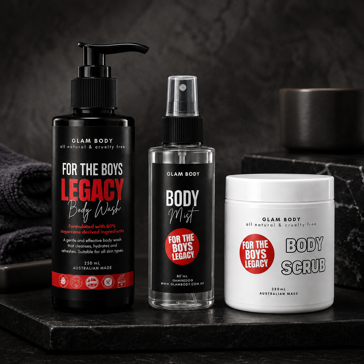 Three Glam Body products arranged on a dark stone surface: a black pump bottle of Legacy Body Wash, a clear spray bottle of Body Mist, and a white tub of Body Scrub. The labels read 'FOR THE BOYS' and show the Glam Body brand.