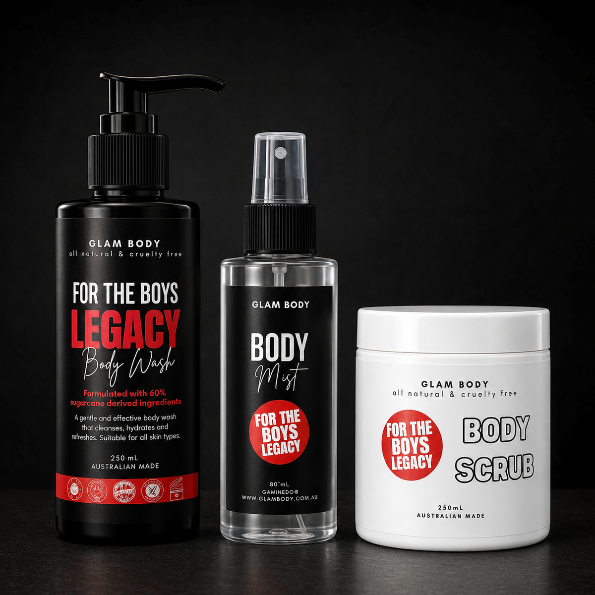 Three body care products from Glam Body are arranged on a dark surface against a black background: a large black pump bottle of Legacy Body Wash, a clear spray bottle of Body Mist, and a white tub of Body Scrub. Each container has black labeling with red and white text.