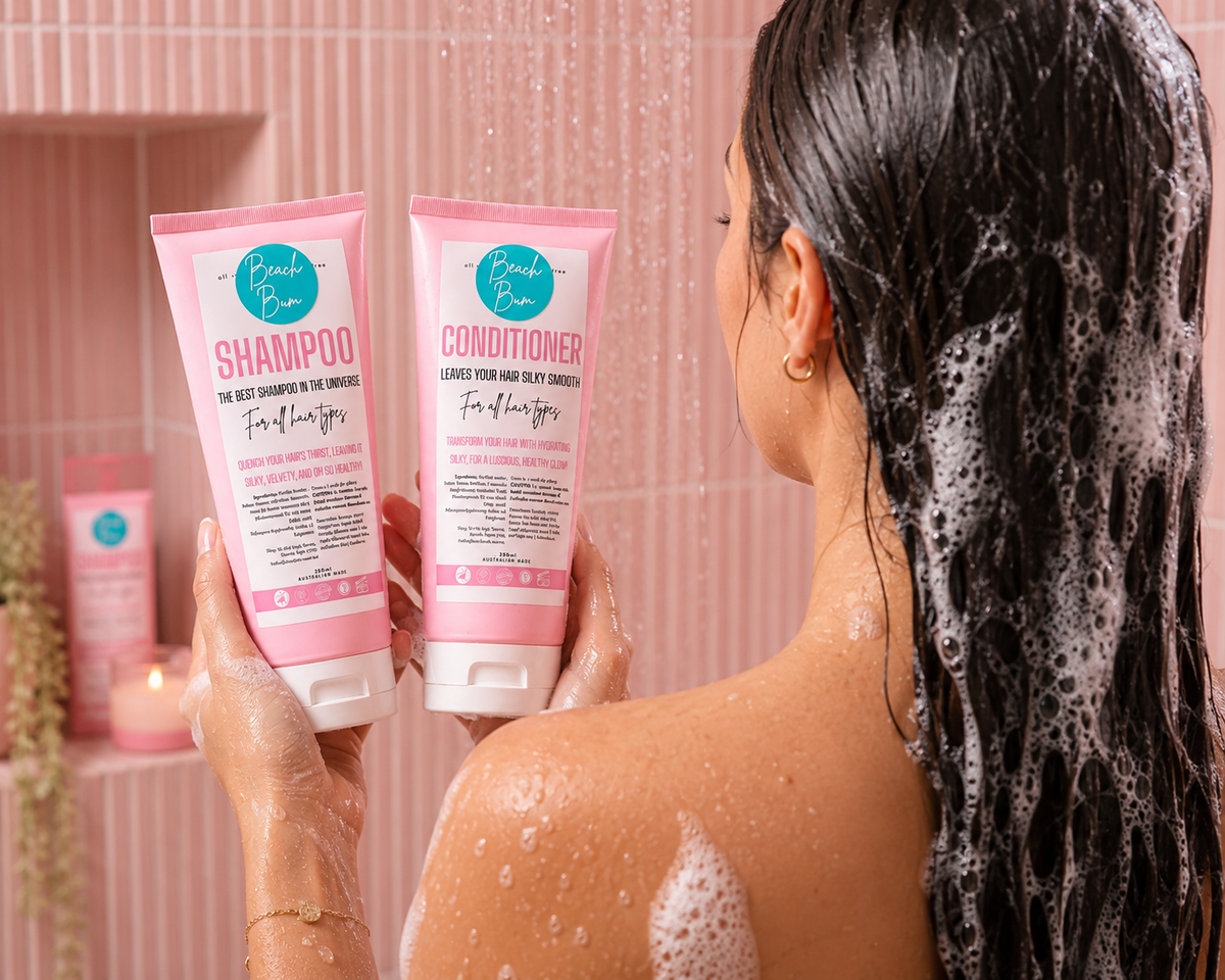 A woman with wet, foamy dark hair stands in a shower holding two pink tubes of hair care products labeled Fresh Pink Shampoo and Conditioner.