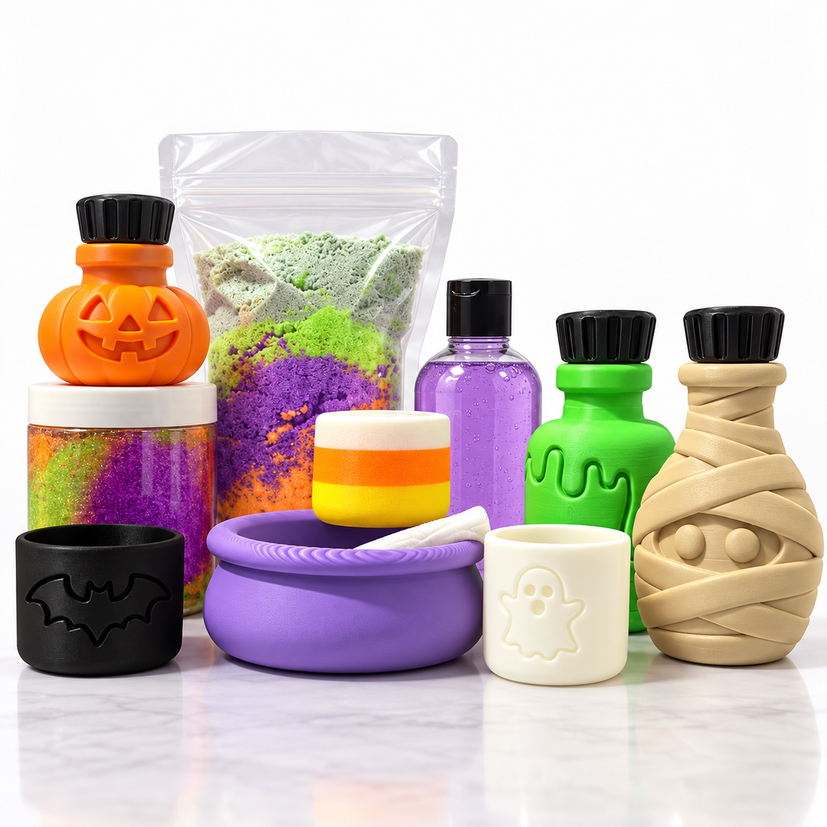 A Halloween-themed bath and body accessory set arranged on a white marble surface against a white background. The set includes a clear bag of multicolor powder, an orange jack-o'-lantern bottle, a purple bottle, a green bottle, a beige mummy bottle, a black bat-shaped ring, a purple bowl, a small striped cup, and a white ghost mug.