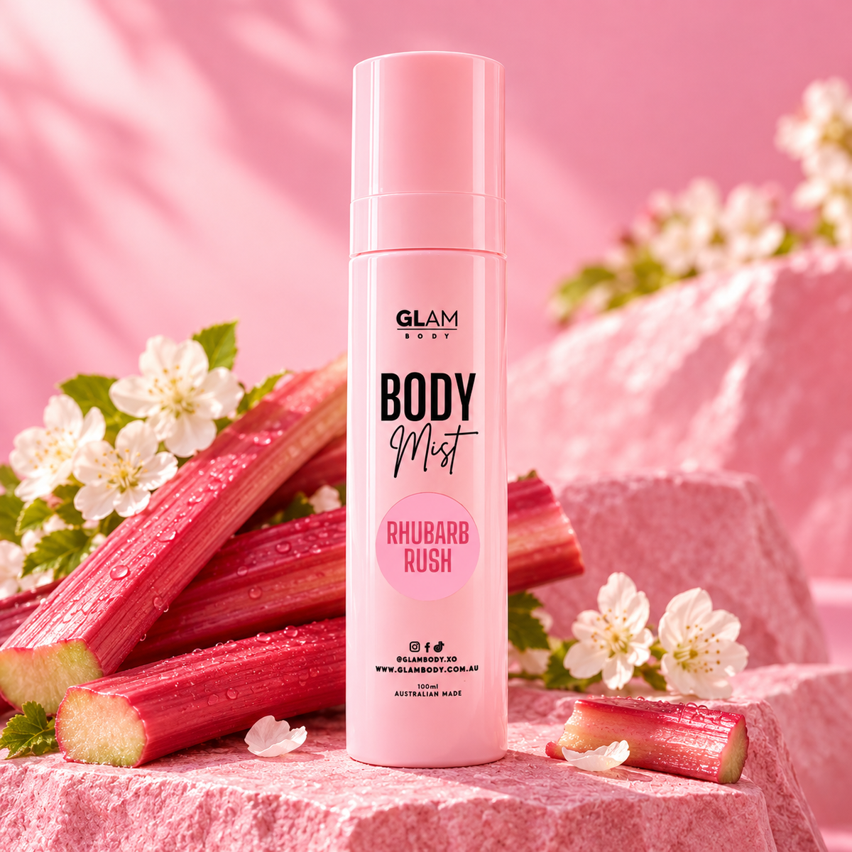 A pink bottle of GLAM BODY Mist, Rhubarb Rush, stands on pink stone blocks surrounded by fresh rhubarb stalks and white blossoms against a soft pink background.