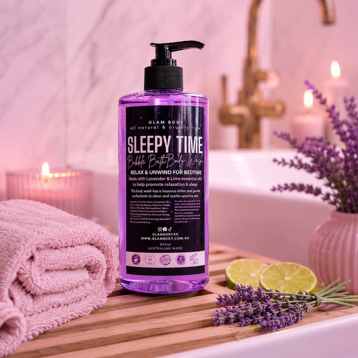 A bottle of Glam Body Sleepy Time Calming Body Wash stands on a wooden tray in a spa-like bathroom setting with lavender, a pink towel, candles, and a pink bathtub in the background.