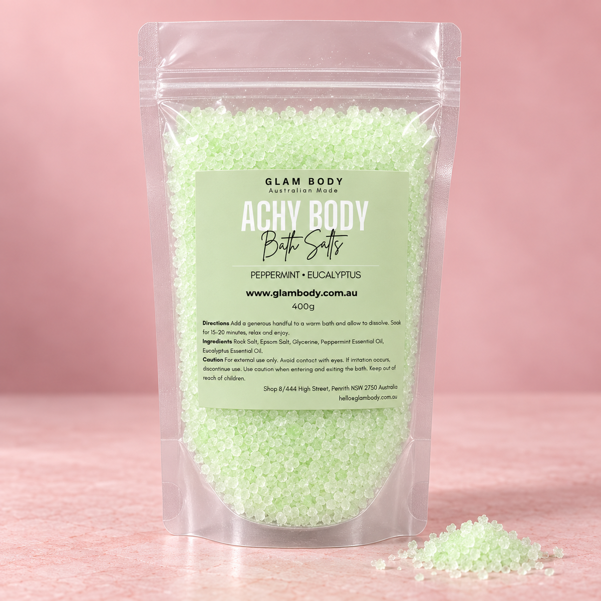 A stand-up pouch of Glam Body Australian Made Achy Body Bath Salts in a peppermint + eucalyptus scent, filled with pale green bath salts, set against a soft pink background with a few salts spilled in front.