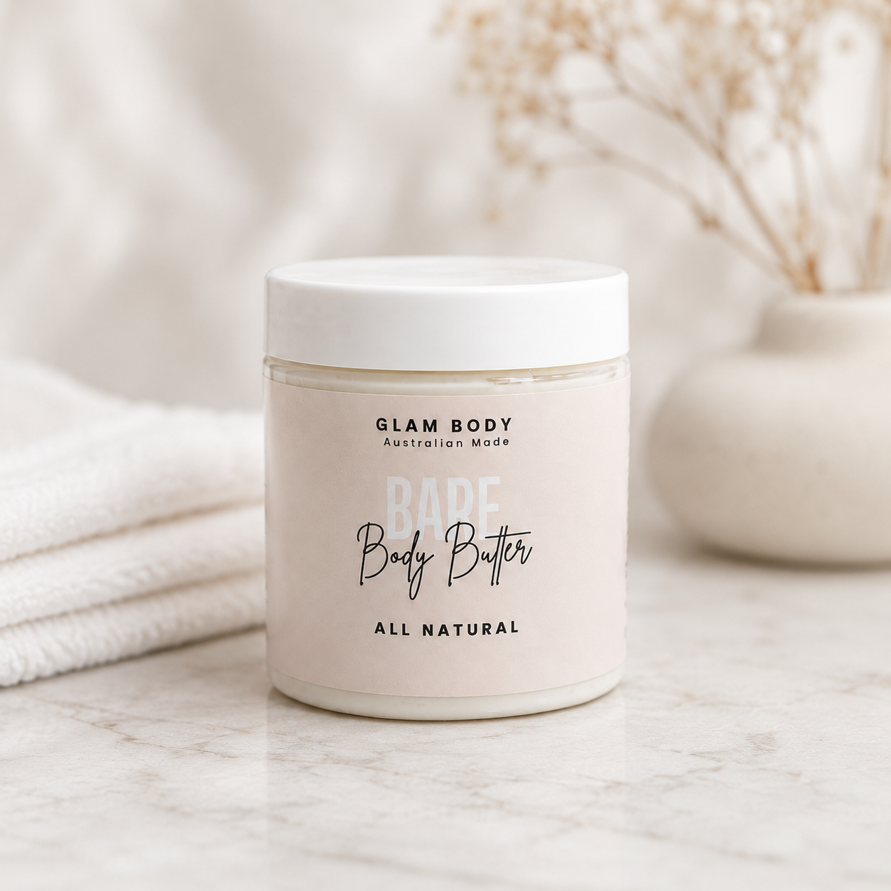 A jar of Glam Body Australia-Made 'RARE Body Butter' with a white lid and a pale pink label, set on a marble surface with a folded white towel and a vase of dried flowers in the background.