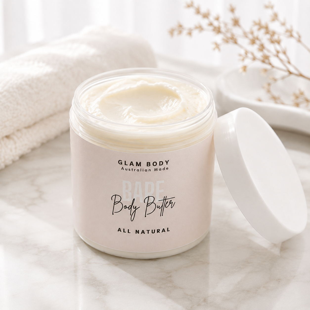 An open jar of Glam Body All Natural Body Butter with its white lid resting beside it on a marble surface, with a soft white towel and dried floral stems in the background.