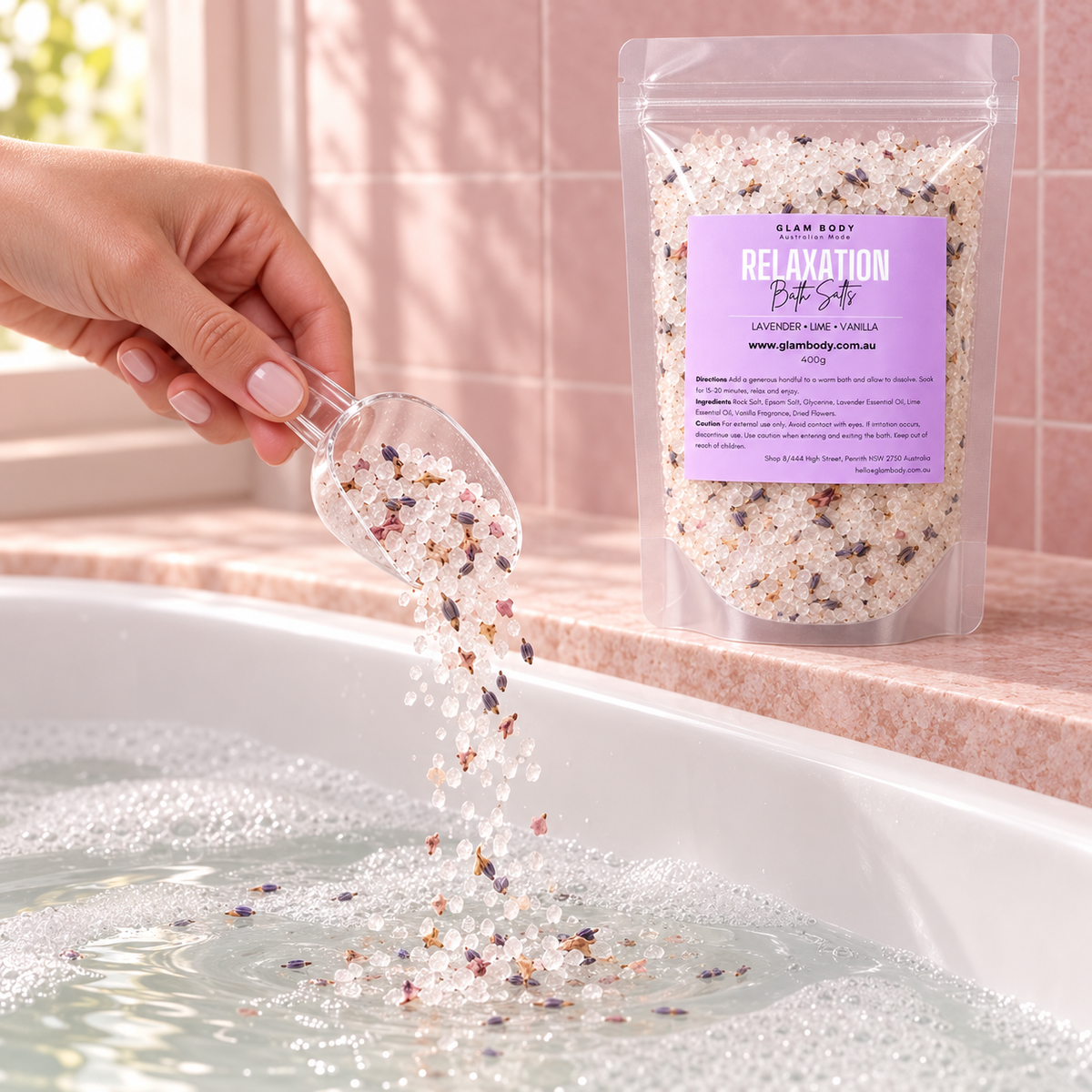 A hand pours colorful bath salts from a clear scoop into a bubbly bathtub, with a stand-up pouch of Slam Body Relaxation Bath Salt standing nearby on a pink tiled ledge.