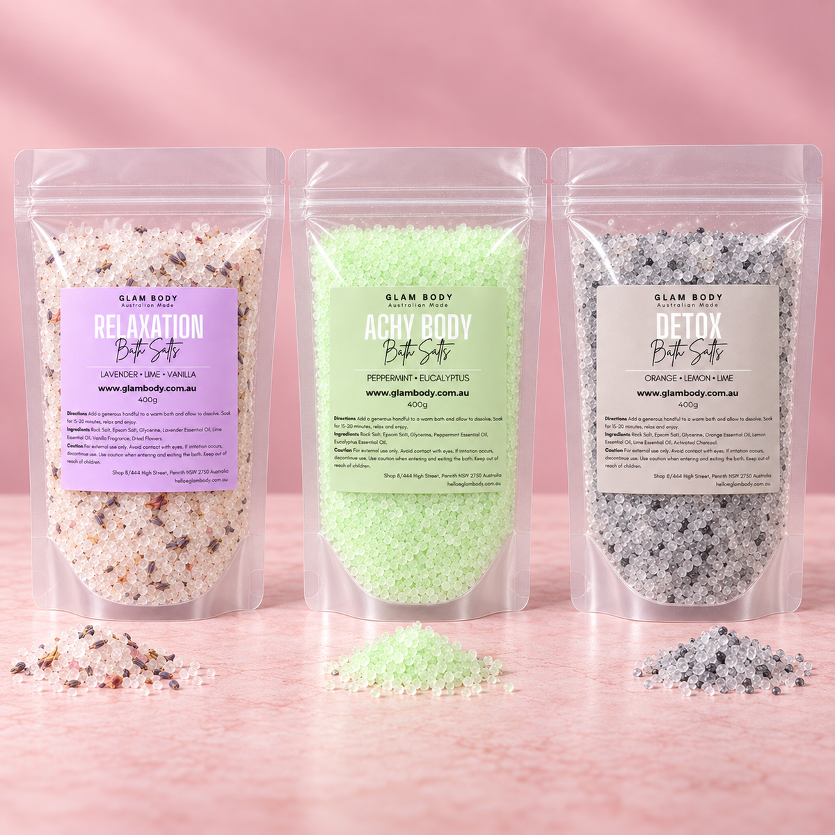 Three stand-up resealable pouches of Glam Body Botanical Bath Salts are arranged on a pink surface against a pink background. The left pouch is lavender-labeled 'Relaxation Bot' with pale pink bath salts inside; the center pouch is mint-green-labeled 'Achy Bot' with light mint bath salts; the right pouch is gray-labeled 'Detox Bot' with dark gray bath salts.