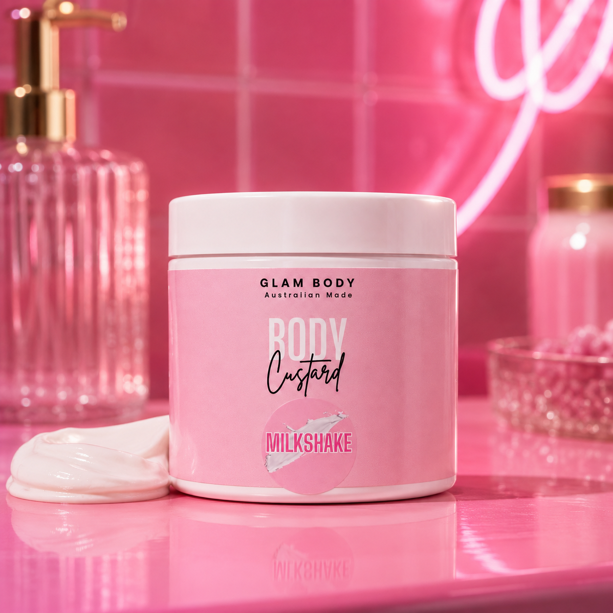 A pink jar of Glam Body Custard in the Milkshake scent sits on a glossy pink surface with a dollop of white cream beside it. The background features a pink tiled wall with a neon sign and blurred pink bath accessories.