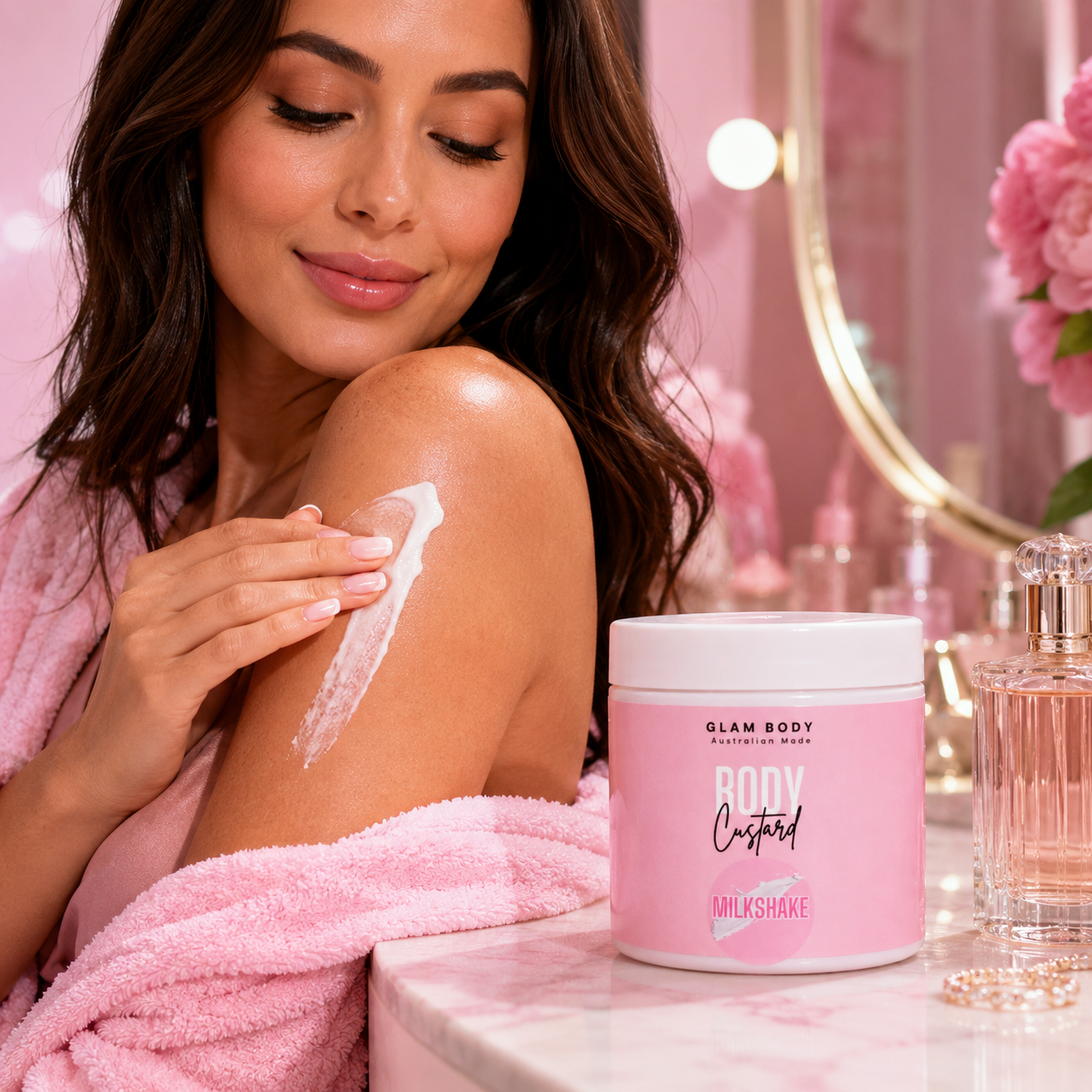 A woman in a pink robe applies white body cream to her upper arm next to a pink tub of Glam Body Milkshake Body Cream on a marble vanity table.