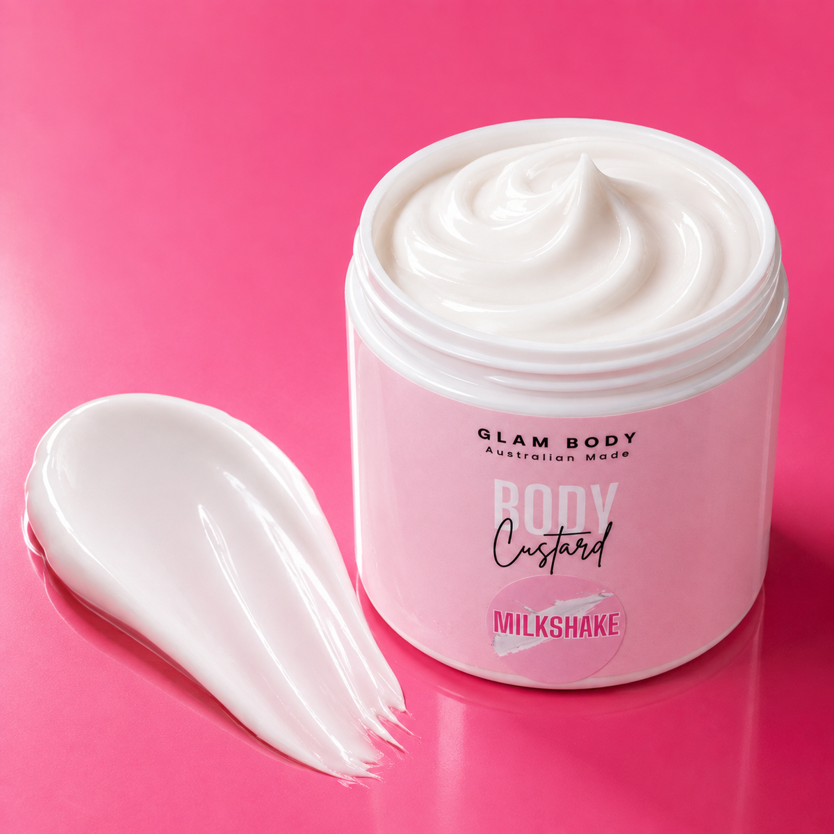 A jar of Glam Body Australian Made Body Custard in the Milkshake flavor, shown open with a swirl of thick white cream inside and a smear of the cream on a vibrant pink background.