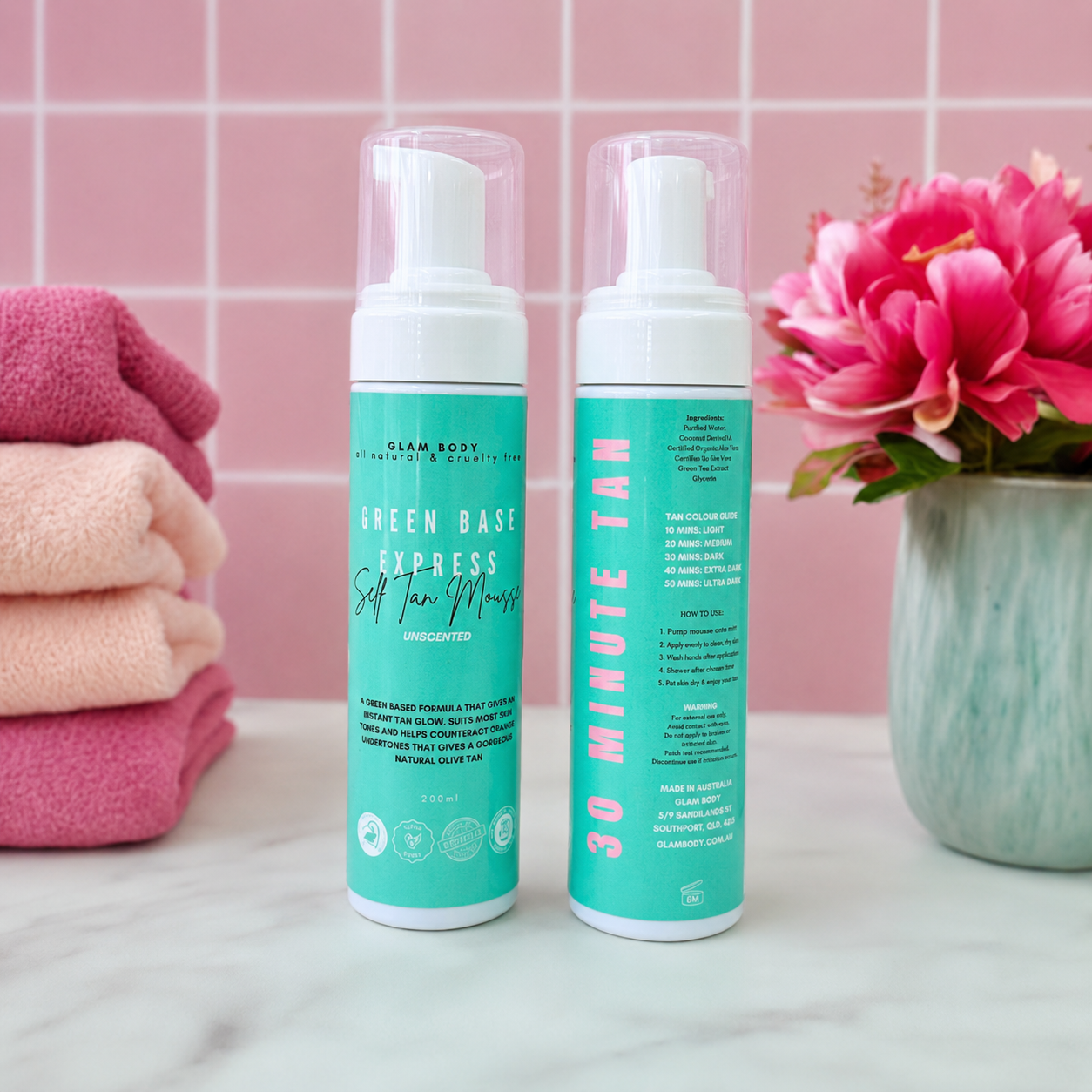 Two cans of glam body Green Base Express Soft Tan Mousse shown on a marble countertop with a pink tiled wall, folded towels, and a pink flower vase in the background.