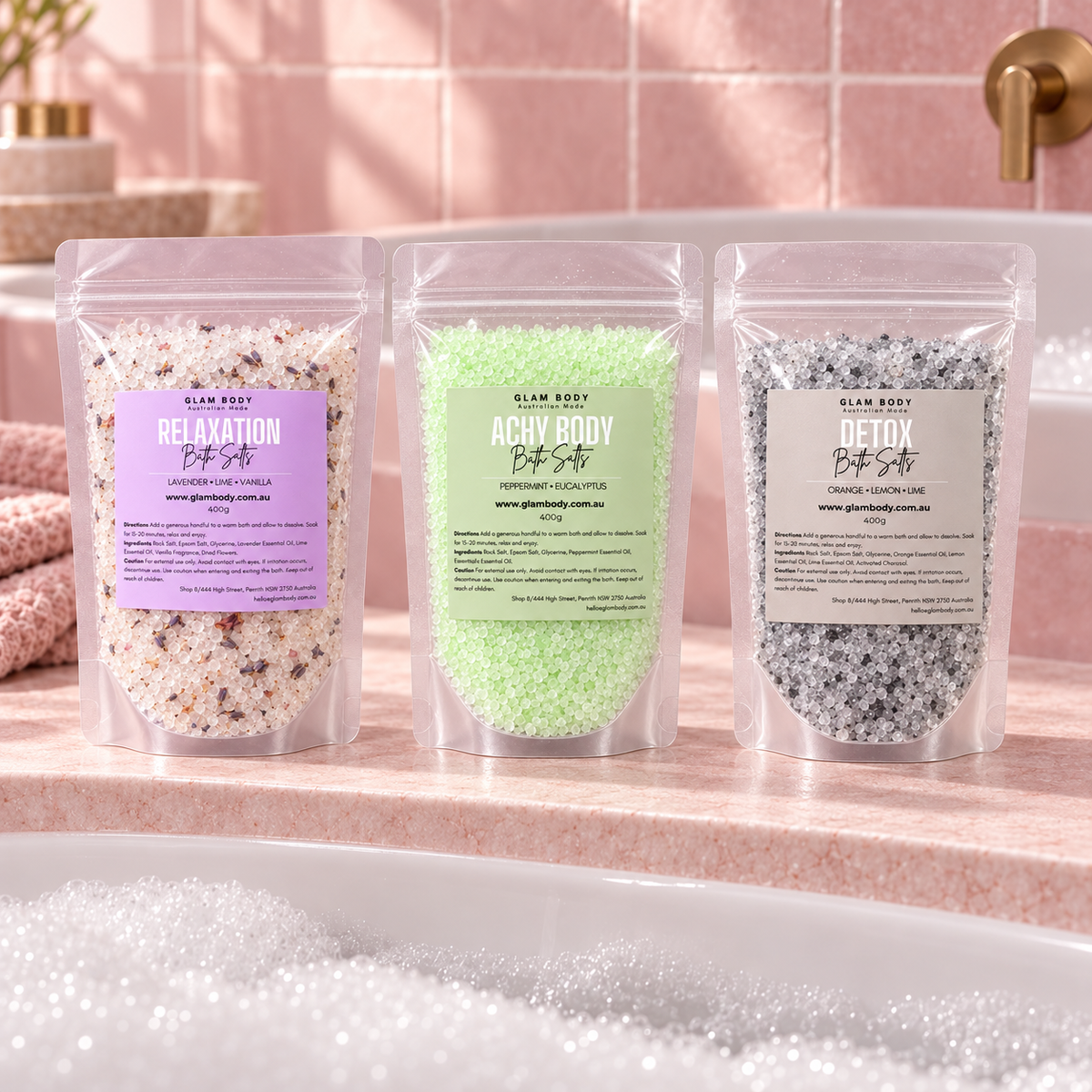 Three stand-up pouches of Glum Body Bath Salt are lined up on a pink marble countertop in front of a bathtub with bubbles. The pouches are labeled Relaxation, Achy Body, and Detox, with lavender, mint green, and gray salt blends visible through the clear packaging.