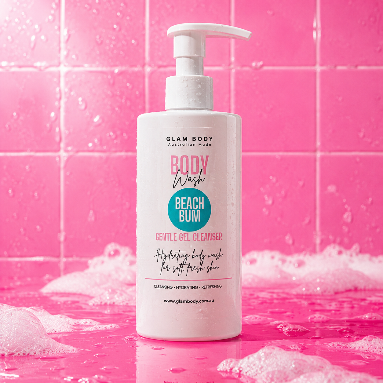 A bottle of Glam Body Beach Bum Body Wash standing upright on a pink tiled surface with pink water droplets and foam around it. The bottle is white with pink and teal text on the label.