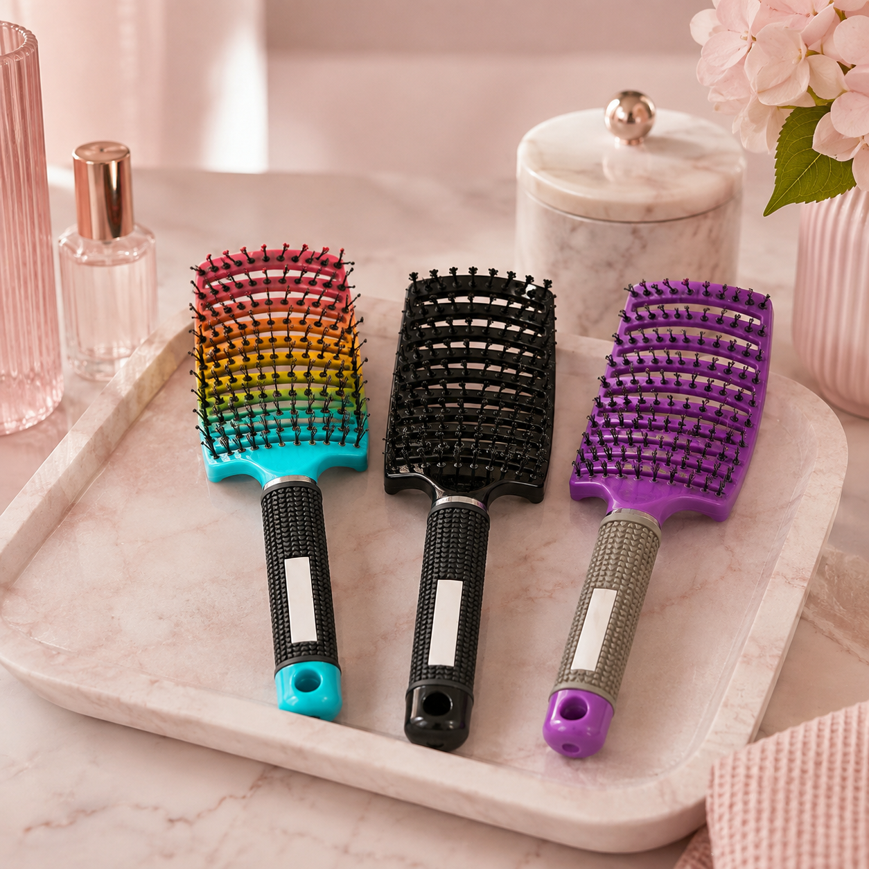 Three wide-tooth hairbrushes arranged on a light marble tray: one rainbow gradient brush with a turquoise handle, one black brush with a black handle, and one purple brush with a gray handle. Pink bathroom-style decor and a pink towel are visible in the background.