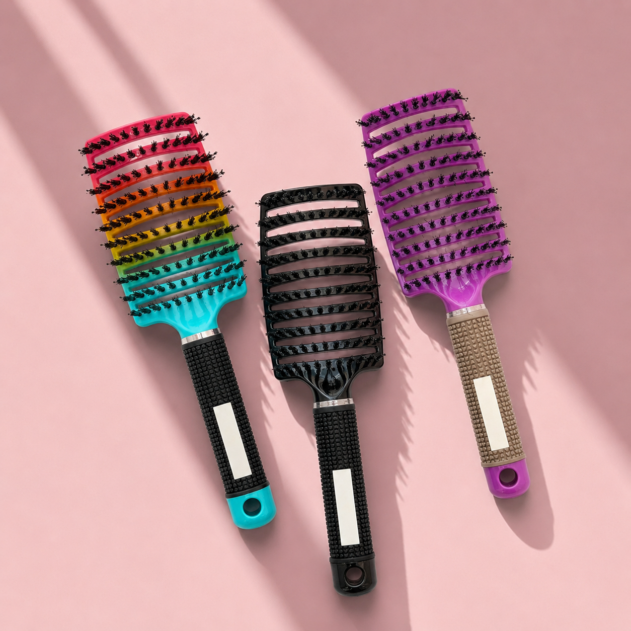Three wide-tooth hairbrushes arranged diagonally on a pink background with soft shadows. The left brush has a rainbow-colored bristle head and a turquoise handle.