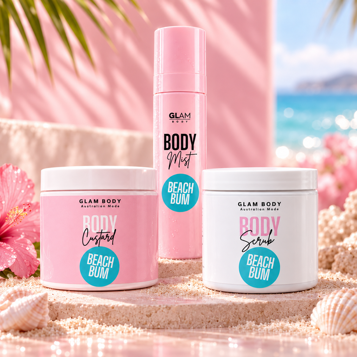 Three Glam Body Australian Made body care products in Beach Bum scent are arranged on a sandy beach-themed set with pink palm leaves, a hibiscus flower, and a blurred ocean background. The set includes a pink spray bottle, a white tub labeled Body Custard, and a white tub labeled Body Scrub.