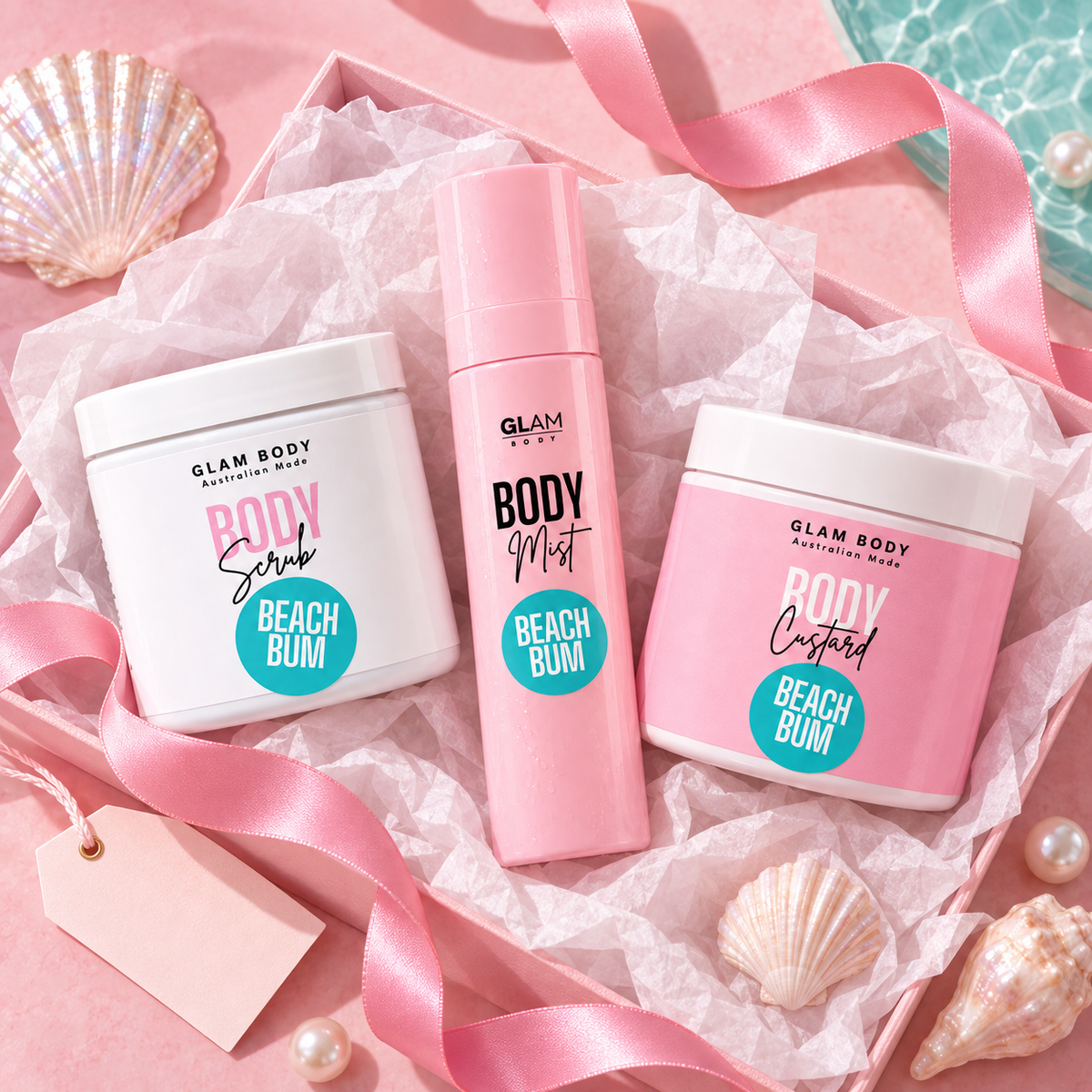 Three Glam Body 'Suitable For Me' body care products in a pink gift box with pink ribbon and seashell props: a white jar of Body Scrub, a pink bottle of Body Mist, and a pink jar of Body Custard, all labeled Beach Bum.