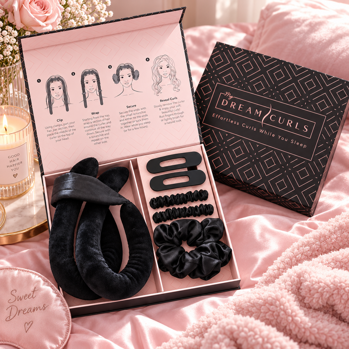 A hair accessory gift set displayed in an open pink box on pink bedding, alongside the branded black box. The set includes black claw clips, black scrunchies, and a black velvet hair bow, with printed curl instructions visible inside the lid.
