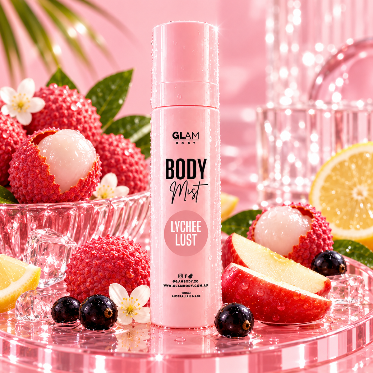 GLAM BODY Body Mist Lychee Lust spray bottle in a pink cylindrical design, surrounded by fresh lychee fruits, sliced red fruit, blueberries, and white flowers on a pink glass tray.