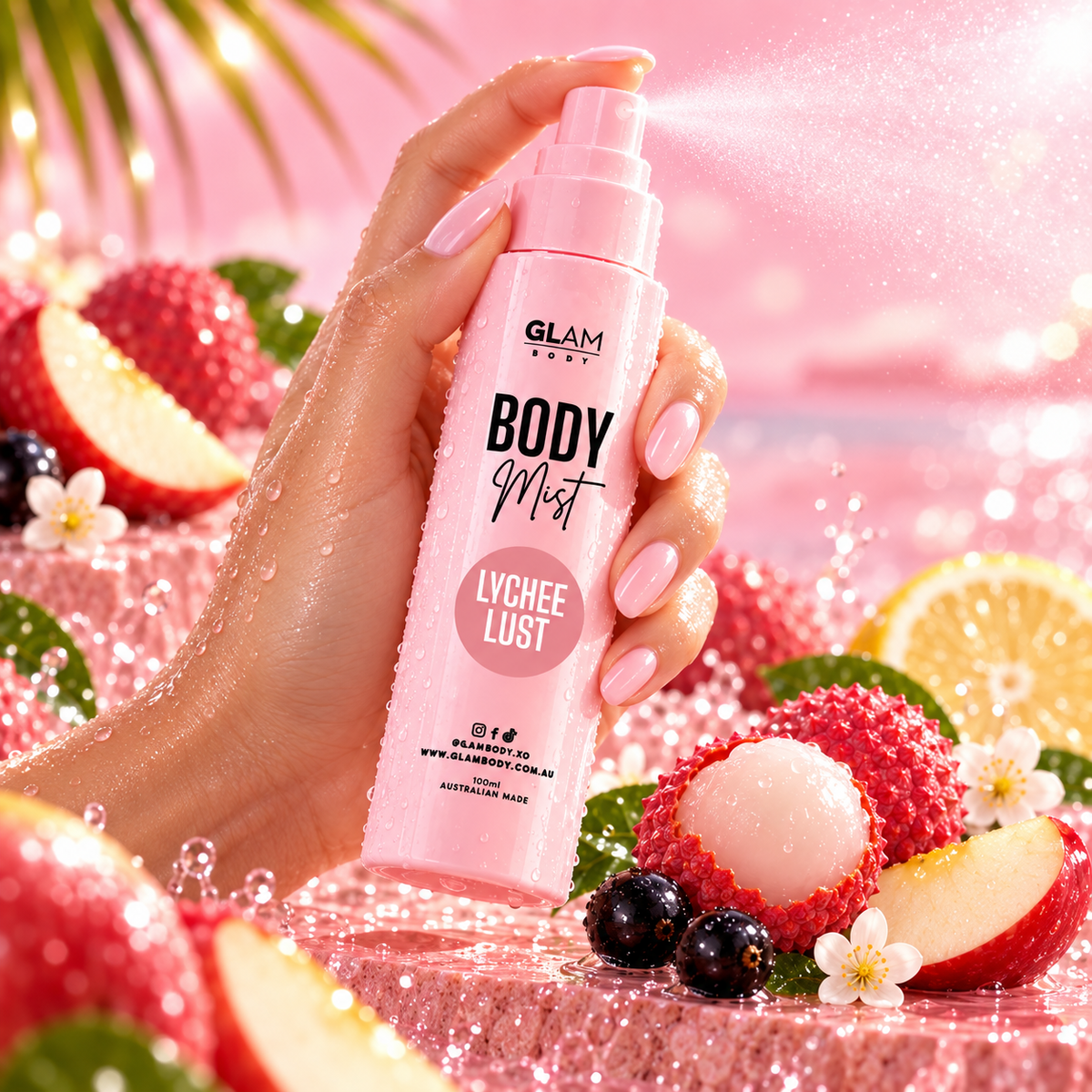 A hand with pink manicured nails holds a pink spray bottle labeled GLAM BEAUTY BODY Mist Lychee Lust, surrounded by fresh lychee fruits, strawberries, blueberries, and white flowers on a sparkling pink background.