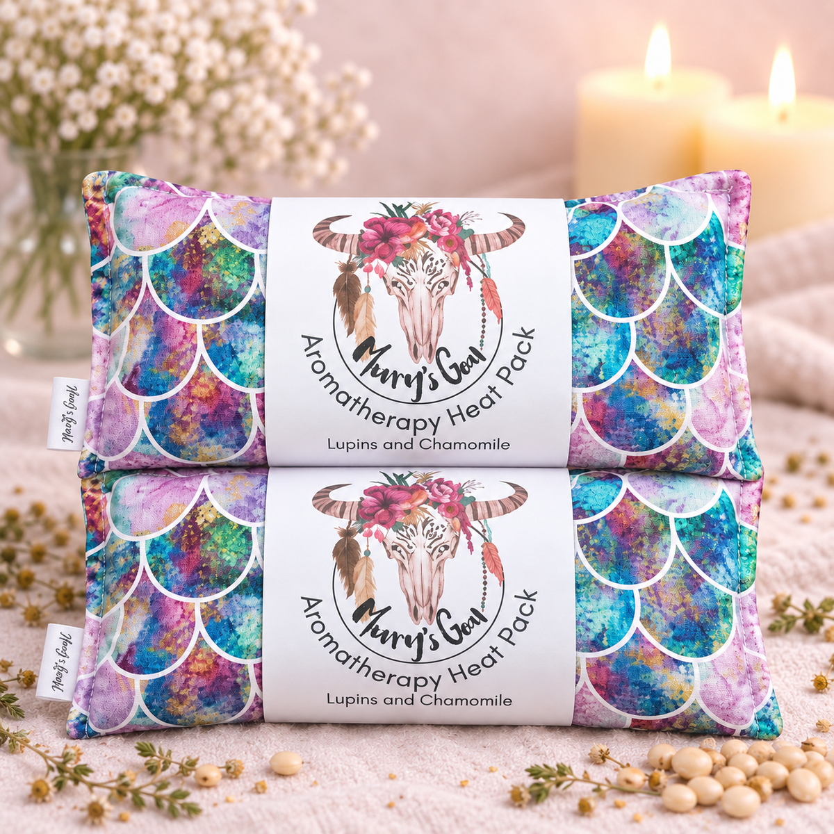 Two 'Merry's Goat' Aromatherapy Heat Packs stacked on top of each other. The packs feature a vibrant, multicolored mermaid scale pattern in shades of blue, purple, pink, and teal, with a white paper band in the center decorated with a watercolor goat illustration and floral accents.