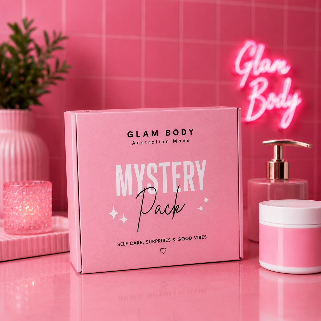 A pink box labeled 'GLAM BODY Australian Made MYSTERY Pack' sits on a glossy pink surface against a pink tiled wall, with a neon sign, candle, plant, and pink cosmetic jar in the background.