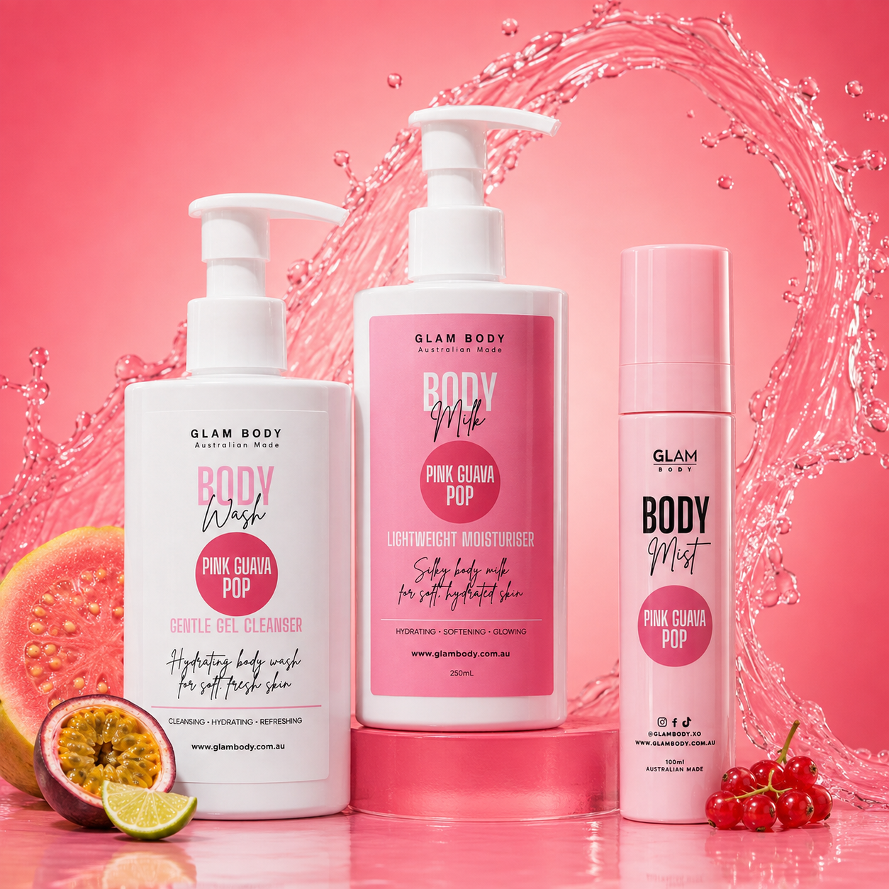Three Glam Body body care products displayed on a pink studio set with fruit props and a water splash backdrop: a white pump bottle of Pink Guana Pop gentle gel cleanser, a white pump bottle of Pink Guana Pop lightweight body moisturizer, and a pink spray bottle of Pink Guana Pop body mist.