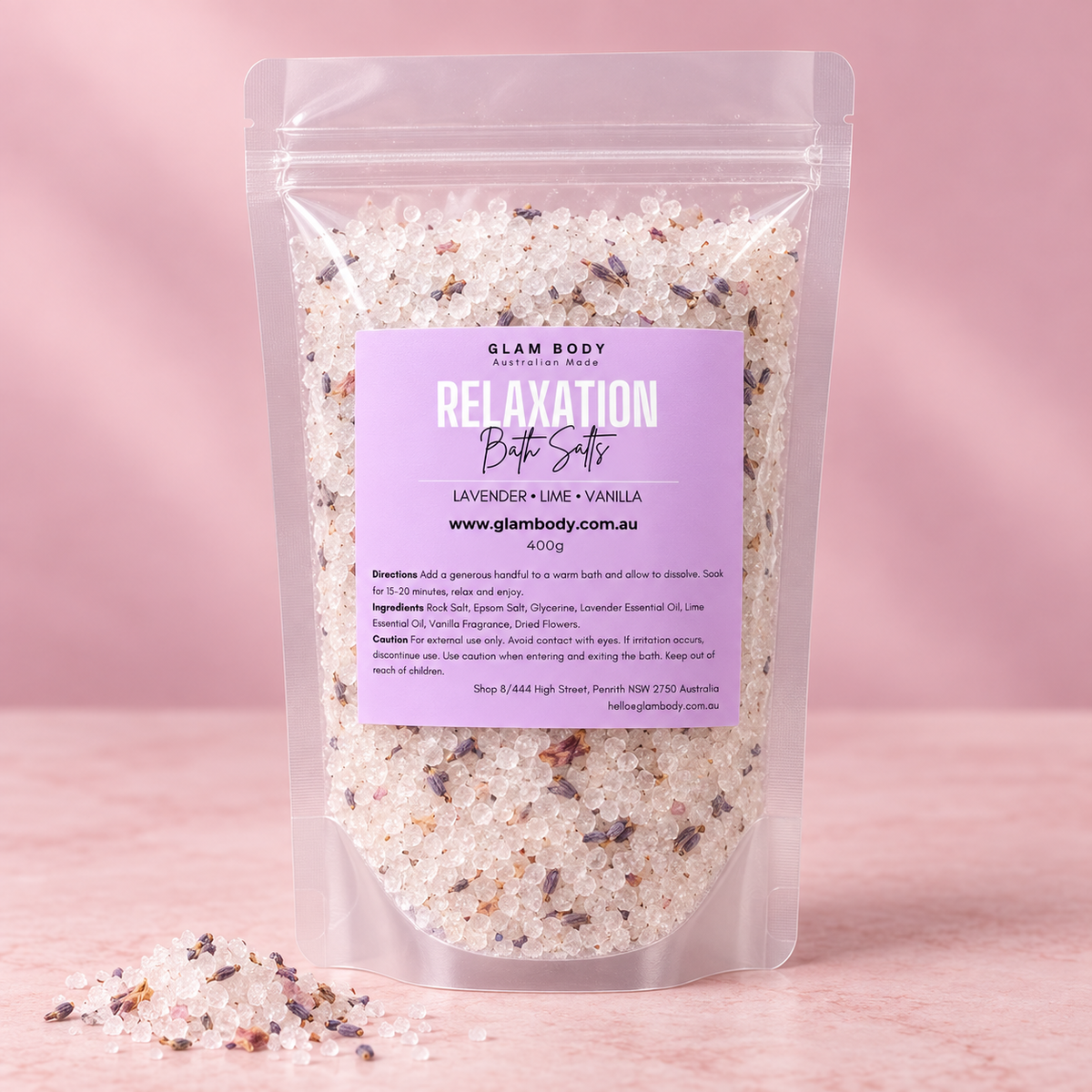 A stand-up resealable pouch of Glam Body Relaxation Bath Salts in lavender and vanilla, filled with white bath salts and dark purple flower pieces, set against a soft pink background with a few salts spilled in front.
