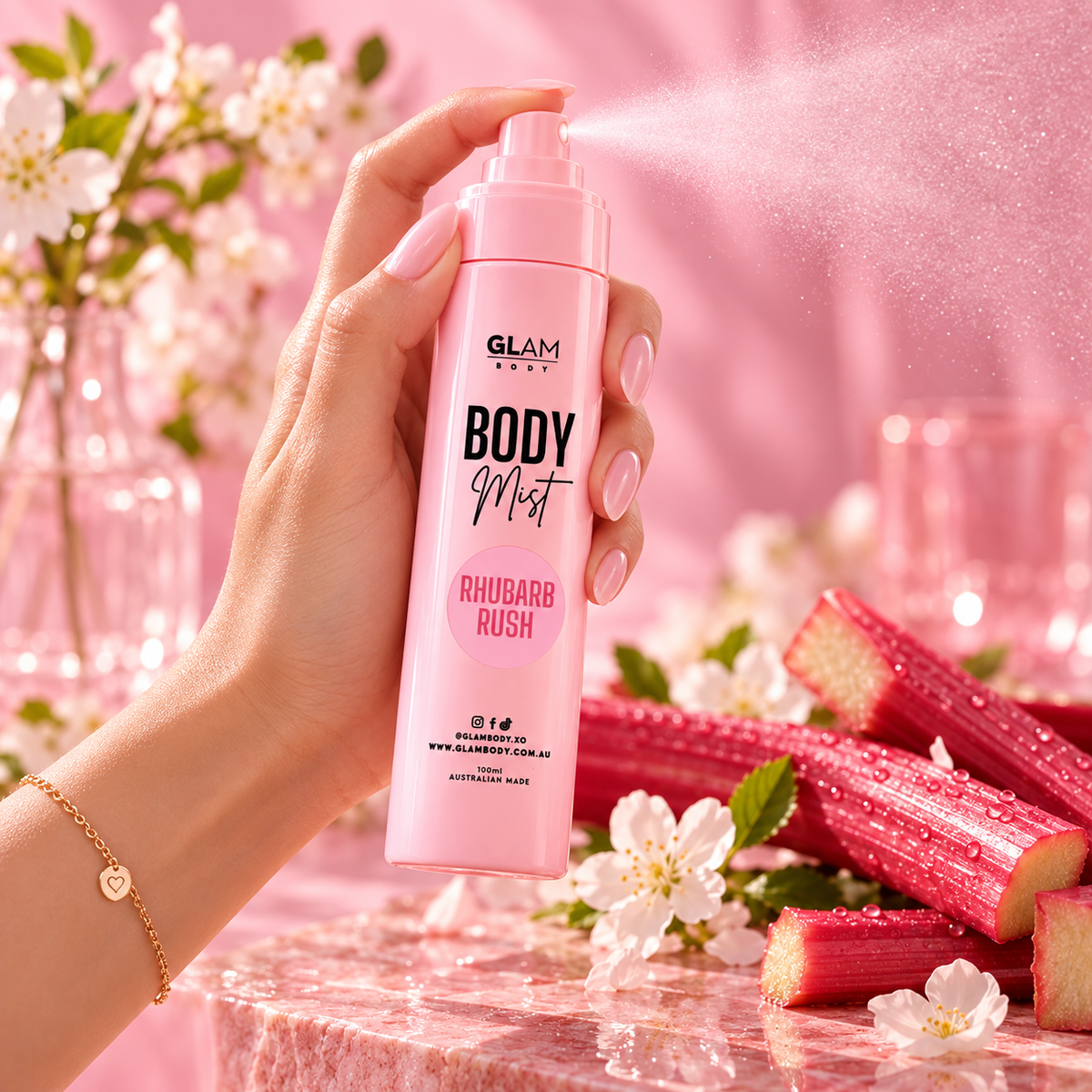 A hand holds a pink Glam Paris Body Mist spray bottle, releasing a fine mist. The background is a soft pink with rhubarb stalks, white flowers, and glassware, creating a romantic, spring-themed aesthetic.