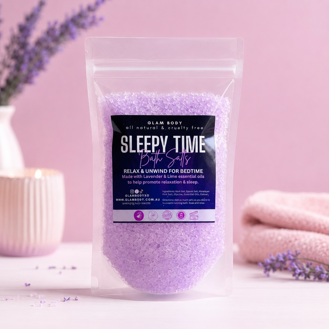 A clear stand-up pouch filled with lavender bath salts, labeled 'Sleepy Time Bath Salts' by Olam Body, set against a soft pink background with decorative lavender and a candle.