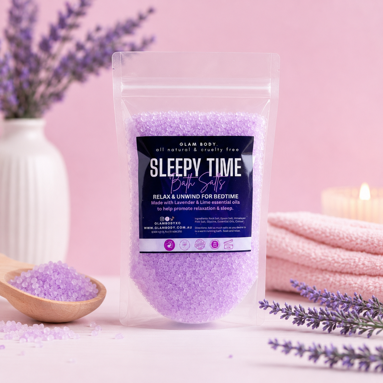 A stand-up pouch of Glam Body Sleepy Time Bath Salts filled with lavender-colored crystals, styled on a pink surface with lavender sprigs, a wooden spoon of bath salts, folded pink towels, and a lit candle in the background.