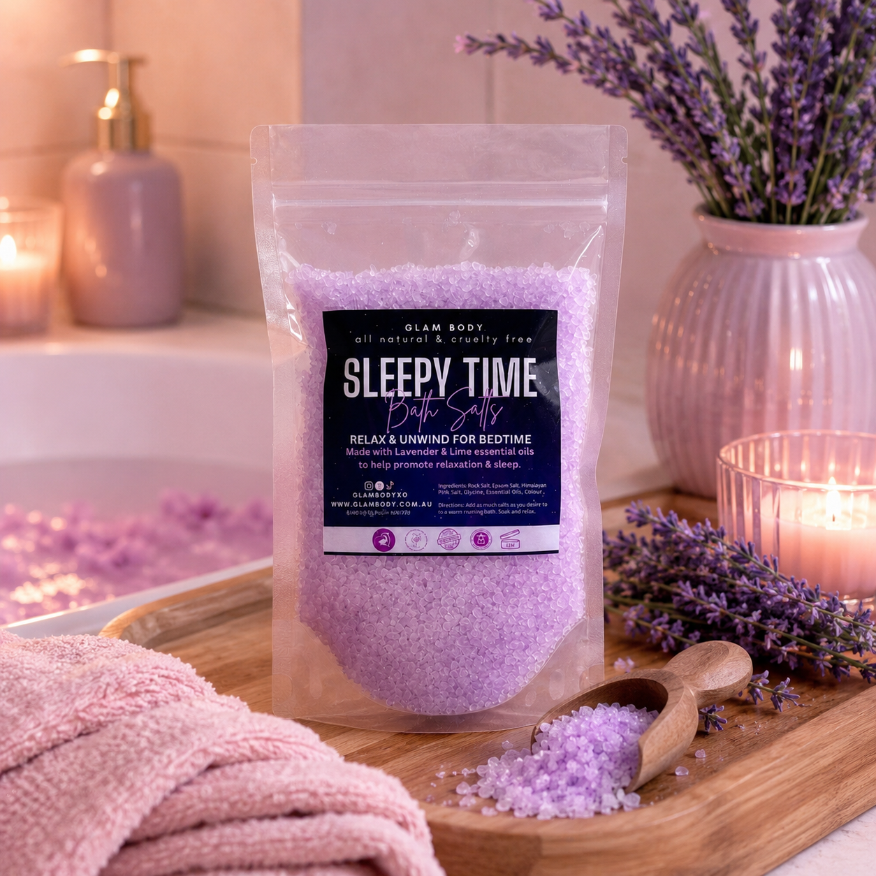 A clear stand-up pouch of Slam Body 'Sleepy Time' bath salts filled with lavender-colored salt, displayed on a wooden tray with a wooden scoop. The scene is styled with a pink towel, lit candles, and lavender sprigs in a warm, cozy bathroom setting.