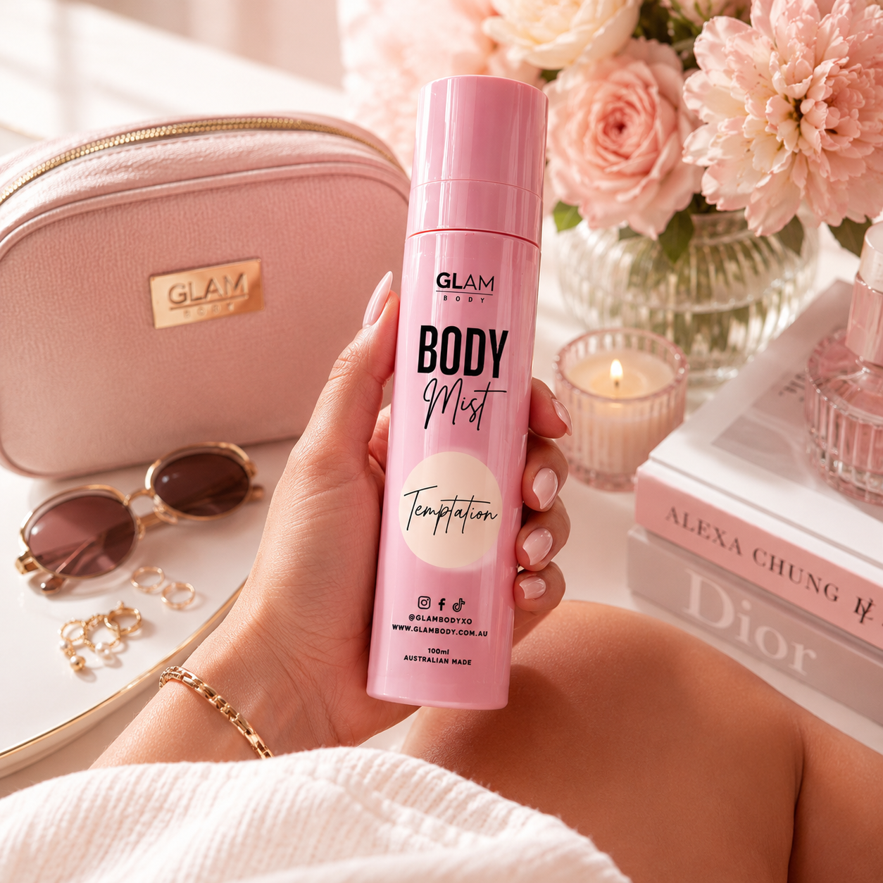Hand holding a pink 'Glam Body' body mist with a blurred background featuring a pink handbag, sunglasses, and flowers.